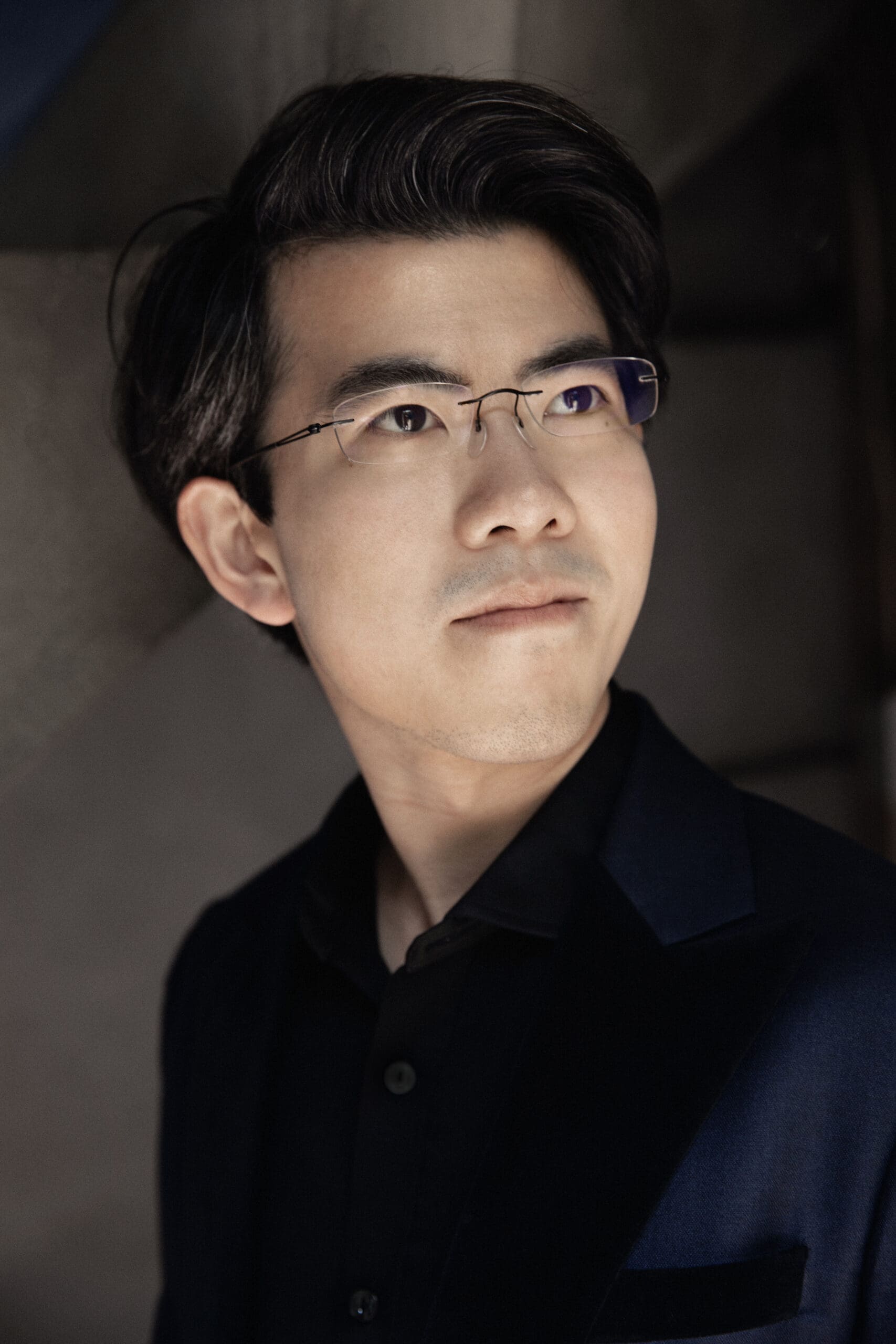 Close-up portrait of a man with dark hair and glasses looking slightly upward.