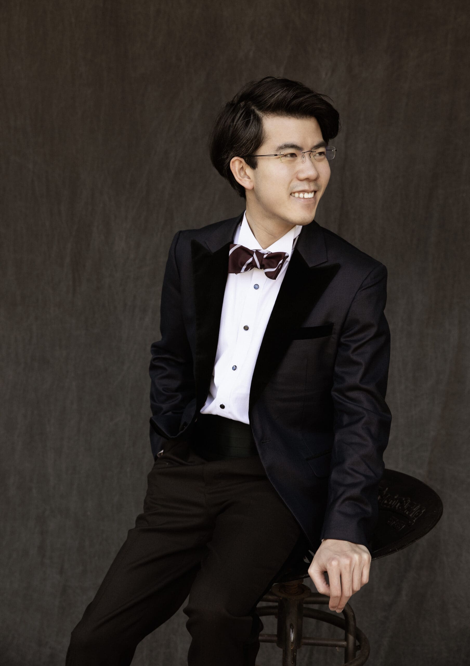 Young man in a black tuxedo with burgundy bow tie, seated on a wooden stool, smiling and looking to the right, holding a top hat in front of him.