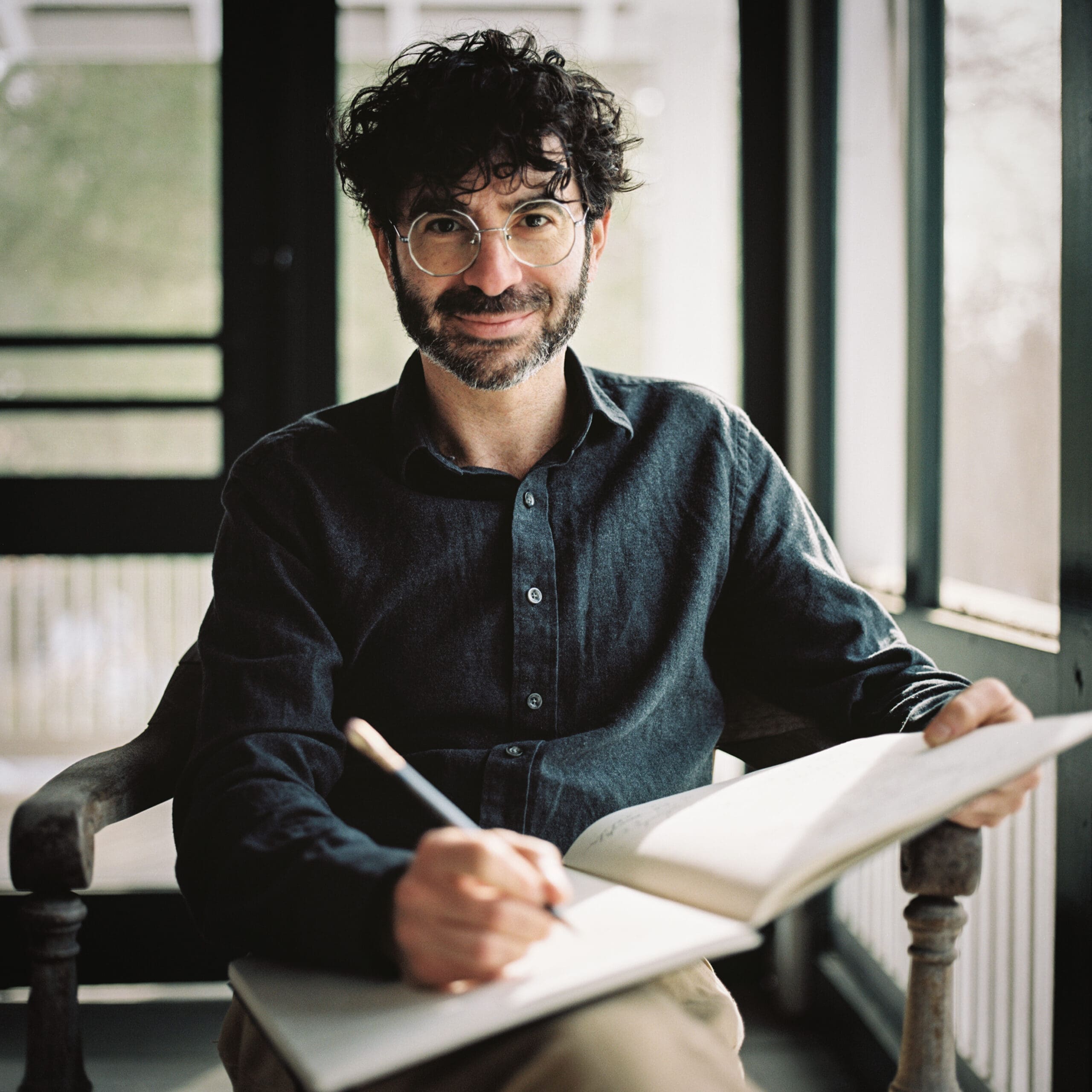 Man with dark, curly hair and round glasses writes in a notebook at a light desk, wearing a dark button-down shirt.