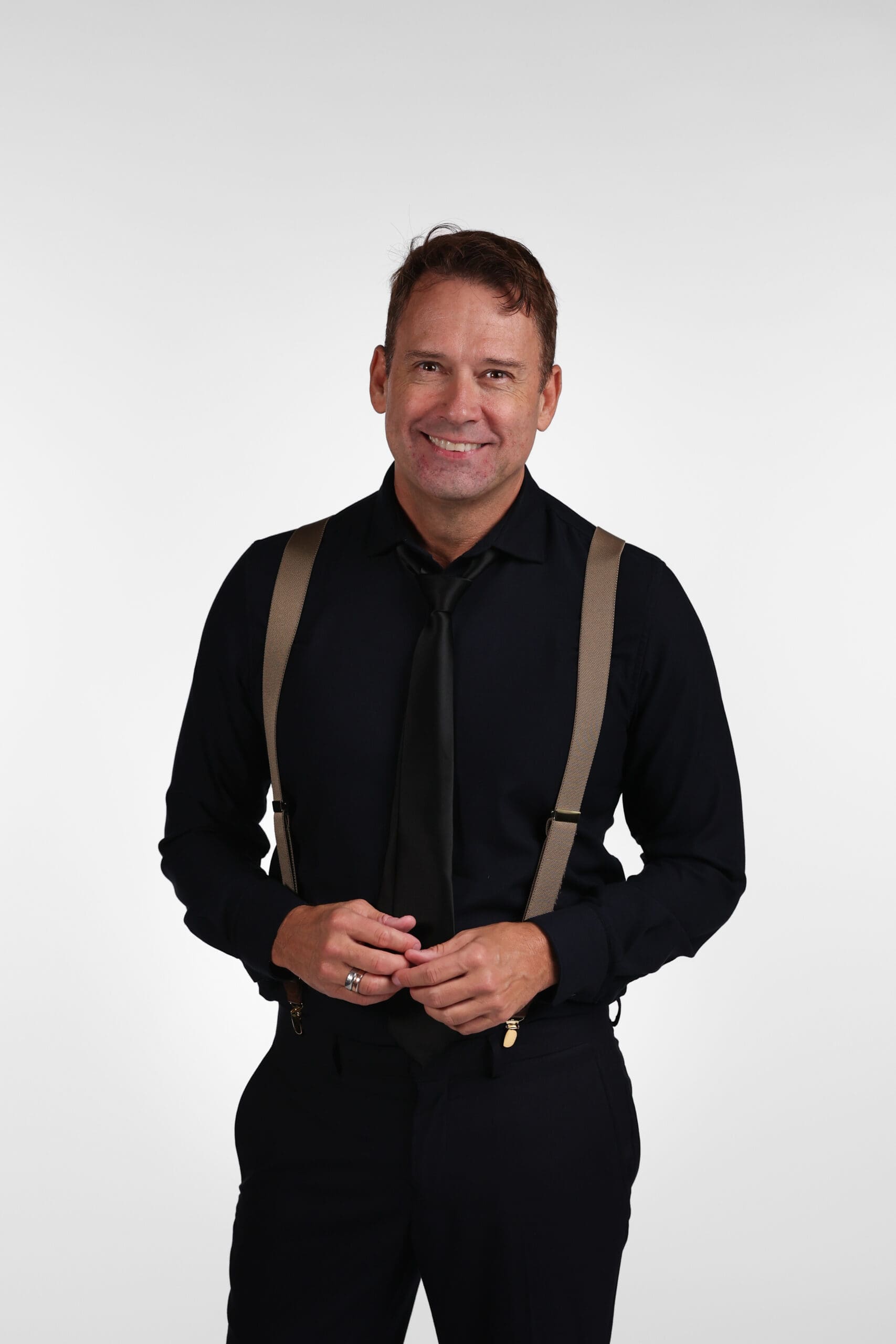 Smiling man in a black shirt with tan suspenders standing against a light background, posing for a portrait.
