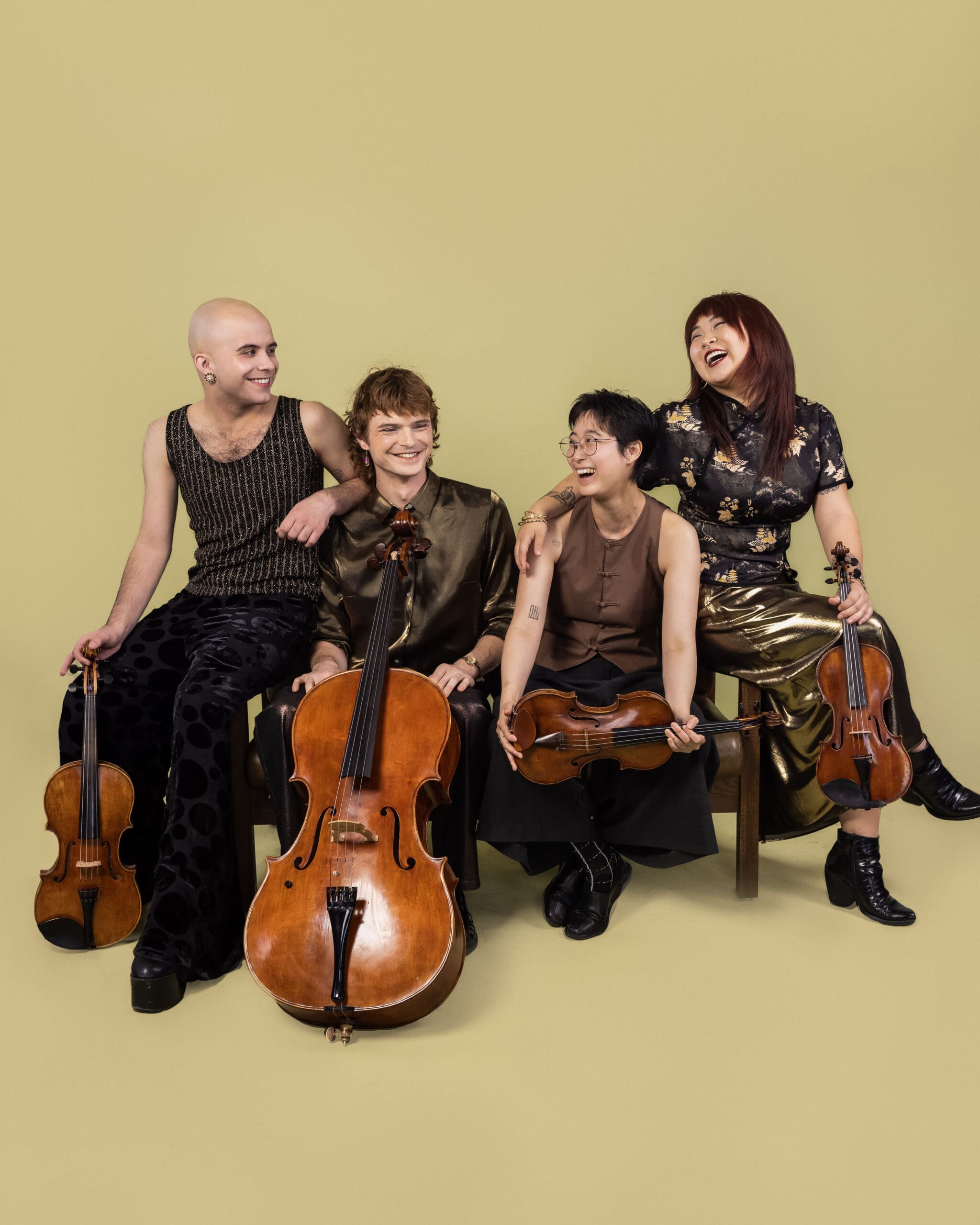 Four diverse musicians sit against a pale yellow backdrop, smiling with their string instruments (cellos and a violin).